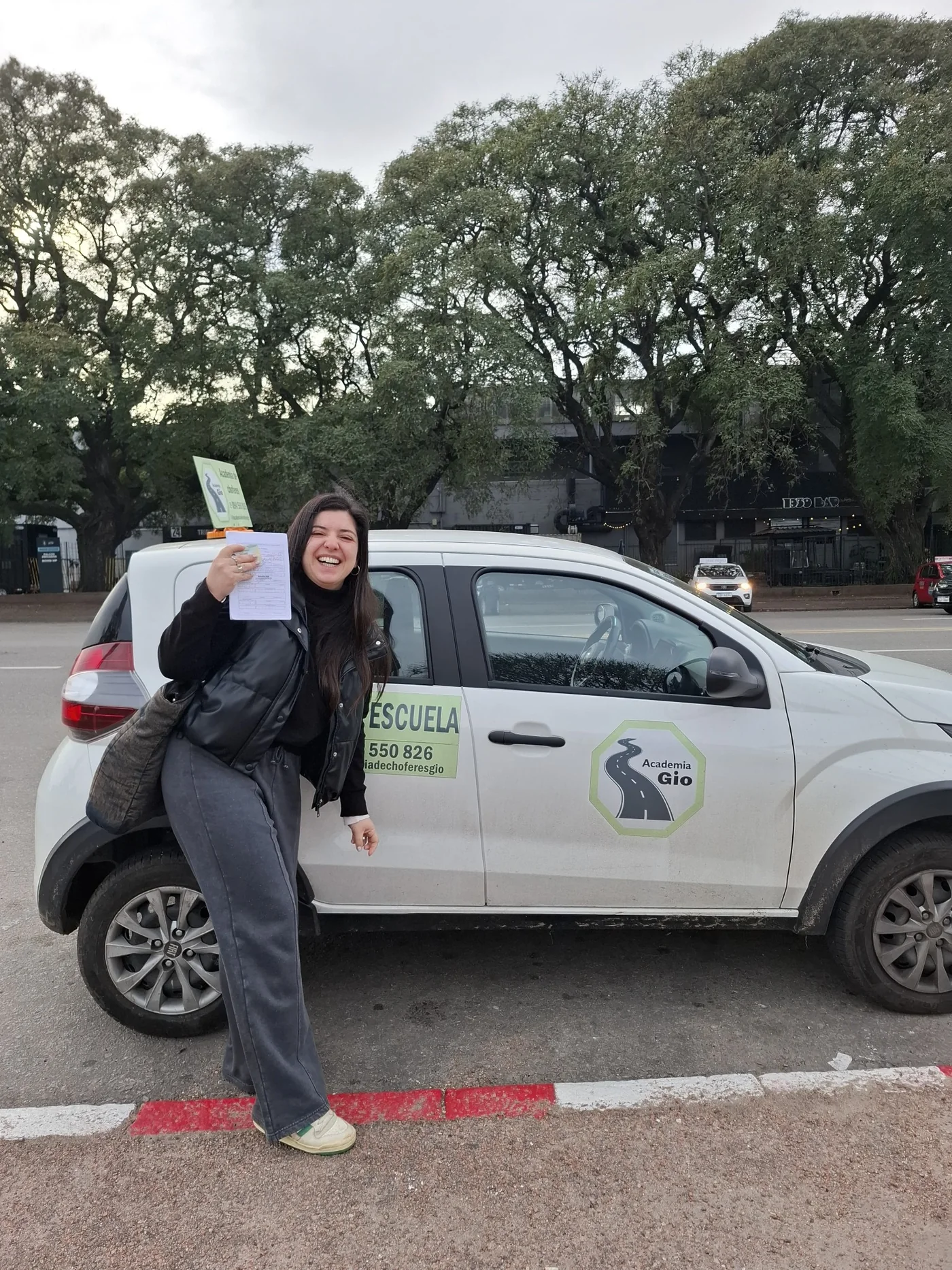 Alumna feliz mostrando la libreta junto al vehículo